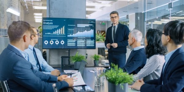 Network Young Stock Trader Shows to the Executive Managers Cryptocurrency and Trade Market Correlation Pointing at the Wall TV.